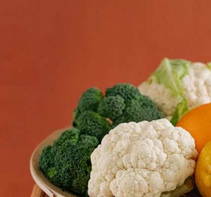 Close-up of fresh broccoli, cauliflower, lemon, and orange on a wooden plate with an earthy background.
